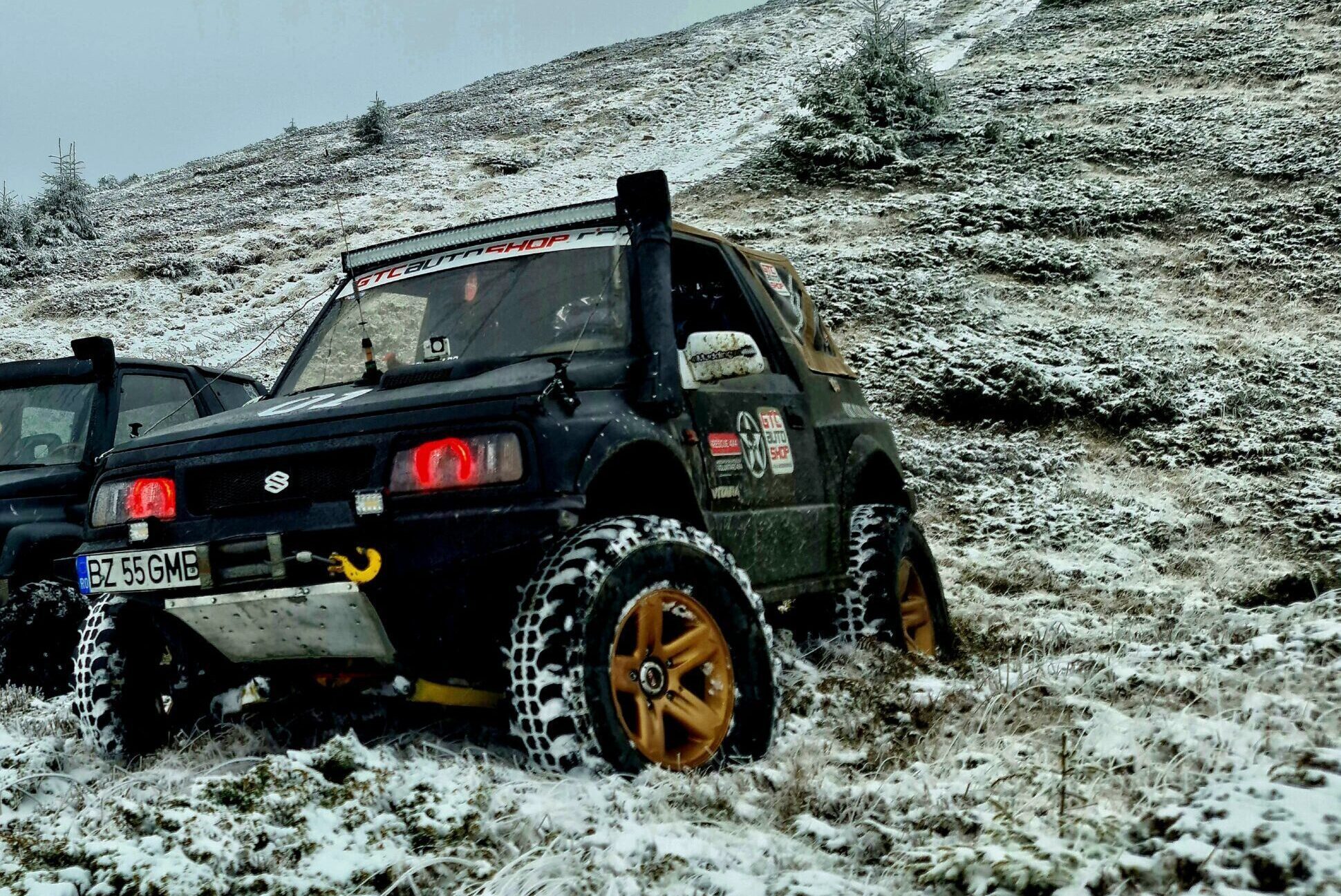 Off-road vehicle on a snowy hillside near a rustic house under a cloudy sky.