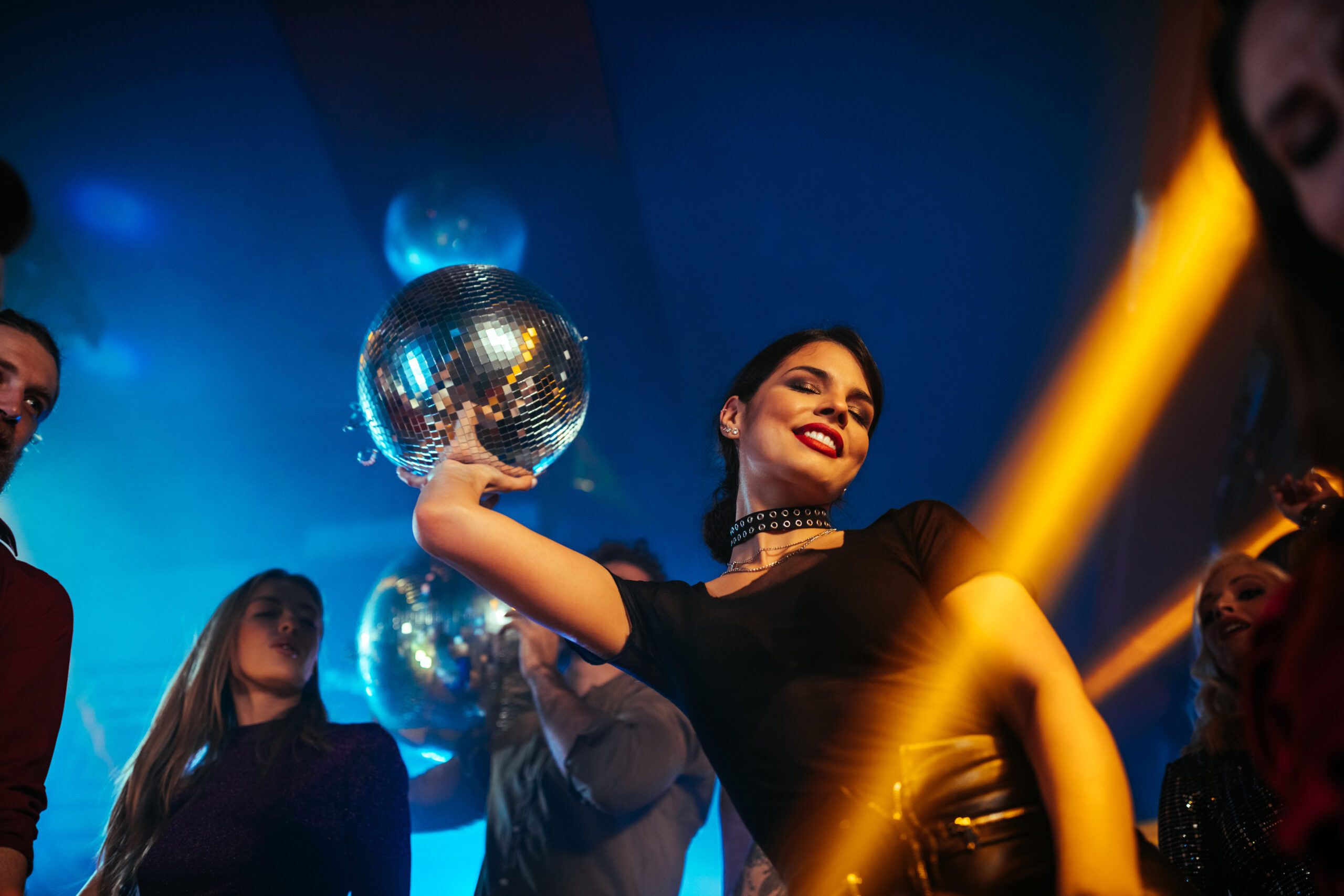 Woman dancing with disco ball at vibrant nightclub party, colorful lights, smiling, lively atmosphere.