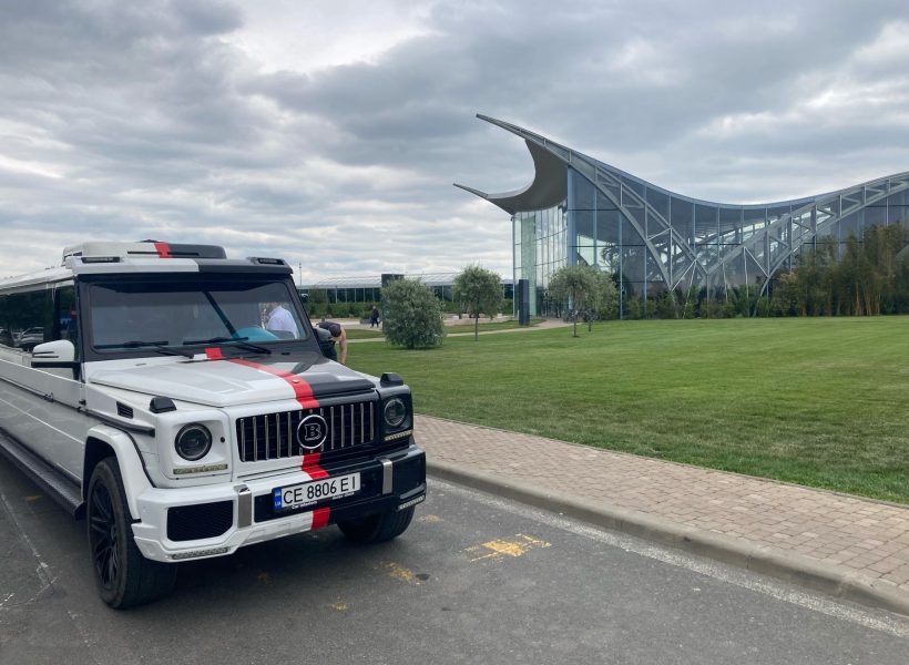 White luxury SUV with black and red stripe parked near modern glass building on a cloudy day.